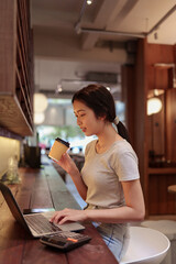 Young woman drinking coffee while working on laptop at stylish cafe counter with warm lighting