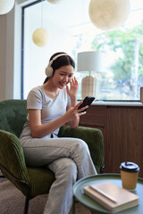 Obraz premium Smiling young woman listening to music with wireless headphones while holding smartphone in cafe setting