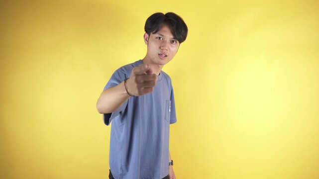 Footage of a Young Asian man in a blue t-shirt against a yellow background, pointing at the camera while grumbling and getting angry