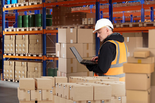 Warehouse employee. Man is storekeeper with laptop. Warehouse employee among boxes. Racks with parcels behind storekeeper. Man in orange vest stands in storage. Warehouse engineer in storage area