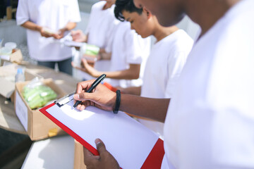 Obraz premium Male Volunteer Checking List on Clipboard While Others Sorting Out Food and Clothes in Donation Center.