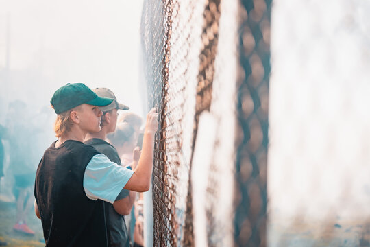 Teenagers watching vehicle burnout competition through chain mesh fence with smoke haze from burning