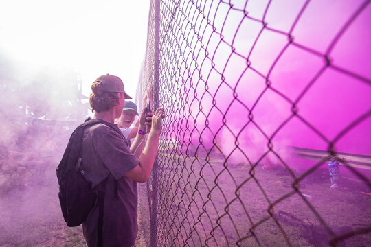 Teenagers watching vehicle burnout competition through chain mesh fence with smoke haze from burning