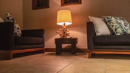 A place to relax in the room. Two sofas with soft patterned cushions against a beige wall in the corner. The desk lamp is lit. Translucent lampshade, designer wooden stand. Evening lighting. 