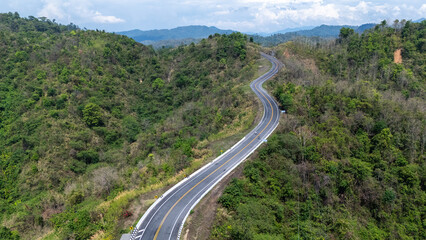 A winding road cuts through lush greenery, surrounded by hills and mountains under a bright blue sky, showcasing the beauty of nature and landscape.