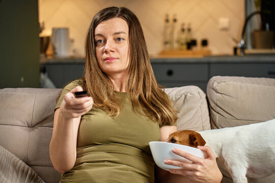 Woman relaxing on sofa with remote control while dog steals food. Pet dog eat food from bowl while owner focuses on TV. Home life and pet behavior concept - Powered by Adobe