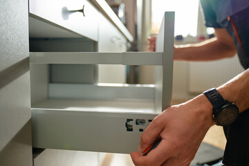 Man assembling kitchen drawer in modern cabinet using screwdriver. Handyman installing and adjusting drawer slide mechanism in contemporary kitchen. Furniture installation