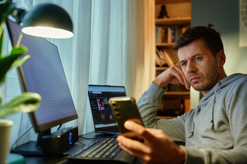 Tired freelancer procrastinating at home office, scrolling phone instead of working. Unproductive man using smartphone at home desk. Concept of procrastination and distraction in remote work