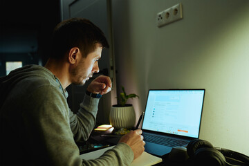 Concentrated freelancer working late at home office. Focused man in hoodie writes notes while working on laptop. Concept of night work