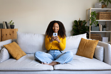 African American woman using mobile phone relaxing at home living room sitting on sofa