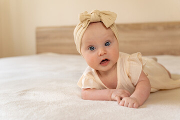 Little baby in a headband and dress lies on the bed at home. Light mocha tones. Fashion for...