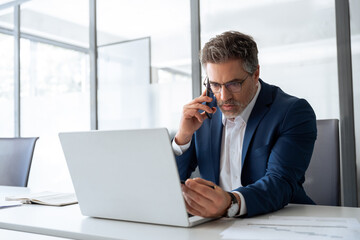 Busy latin senior man trader working on laptop computer in office, having phone call with finance consultation. Focused mature businessman professional using pc, thinking analysing business strategy