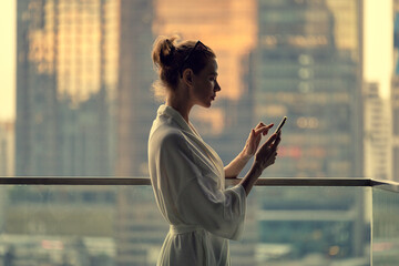 A girl in a dressing gown stands on a balcony and uses a smartphone. An evening metropolis is in the background.