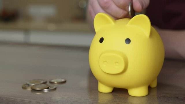 A person adds a coin into a cheerful yellow piggy bank while surrounded by scattered coins. This activity takes place in a cozy home setting, promoting savings and financial habits.