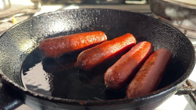 Four sausages are fried until crispy in a black cast-iron old frying pan in golden olive oil, which is already starting to smoke because the sausages have been overcooked in slow motion