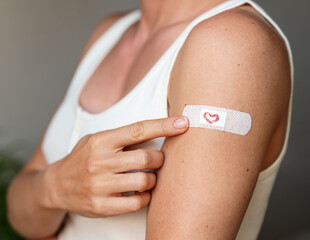 Female pointing at adhesive bandage with drawn heart on her arm at vaccination site.