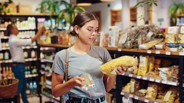 Smiling attractive young girl carefully comparing different types of pasta, deciding which to purchase while standing among grocery shelves in organic food store