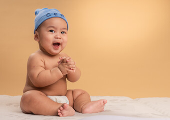 Asian baby wearing a blue “CEO” beanie sits confidently on a mattress with a curious...
