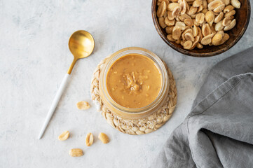Glass jar with creamy peanut butter on grey background with scattered roasted nuts