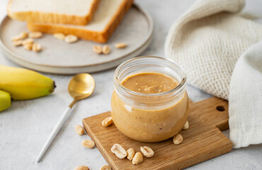 Glass jar filled with creamy peanut butter on grey background with bananas and fresh bread.
