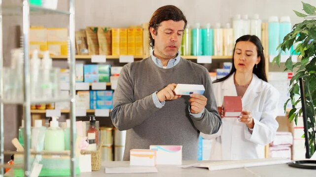 Attentive middle-aged man choosing ointment in box while female pharmacist giving offer in chemistry. High quality 4k footage