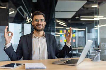 A smiling businessman sits at his desk in the office, gesturing with his hands in a zen-like expression. He appears calm and focused, embracing a moment of mindfulness