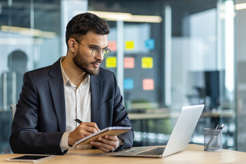 A focused businessman, clad in a suit, meticulously takes notes while seated at a desk, utilizing a...