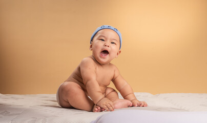 Asian baby wearing a blue “CEO” beanie sits confidently on a mattress with a curious expression. The child is shirtless, wearing a diaper, against a warm beige studio background