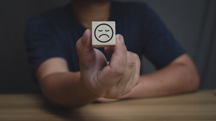 A hand holds a wooden block displaying a sad face emoticon, symbolizing negative emotions, dissatisfaction, and disappointment.