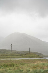 Fototapeta premium Scenic lake in the Connemara region of Ireland, surrounded by lush green hills, cloudy skies, and a traditional stone wall in the foreground.