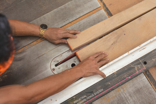 Close-up of hands guiding a wooden board through a table saw with a red blade, used for precise straight cuts in woodworking.