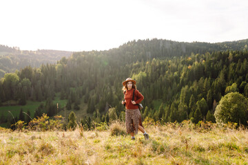 Beautiful female tourist with backpack exploring vibrant green valley. Young woman in hat enjoying sunset and nature. Travel, nature and freedom concept. Active lifestyle.