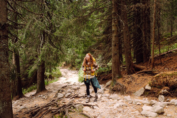 Obraz premium Young female hiker with hiking backpack walking up rocky path in lush green forest. Happy woman hiker enjoying nature on sunny day. Adventure, recreation concept. Active lifestyle.