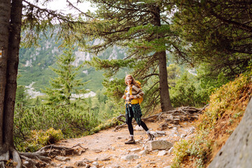 Fototapeta premium Young female hiker with hiking backpack walking up rocky path in lush green forest. Happy woman hiker enjoying nature on sunny day. Adventure, recreation concept. Active lifestyle.