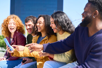 Group of diverse friends laughing together while using smartphone outdoors
