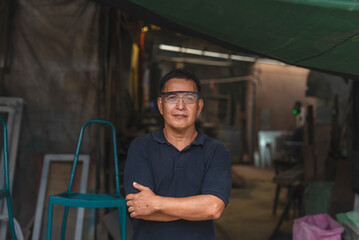Portrait of a seasoned craftsman standing confidently with arms crossed in front of a humble, hands-on furniture workshop.