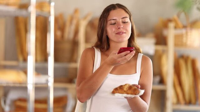 Glad young girl carefully scans croissant, counting calories at the bakery shop