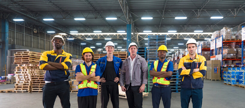 Group of factory labor stand together with arms crossed in industry factory. Everyone wearing safety uniform and helmet. Work atmosphere in general warehouse with racking system and storage facility.