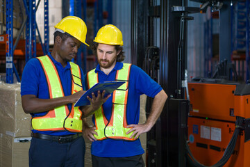 Two warehouse worker in yellow hard hat and safety vest discuss operational detail while examining document in an industrial setting. They focus intently on their task.