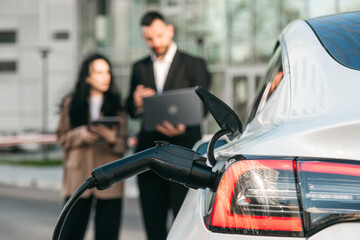 A business man and woman work on a laptop while their electric car charges in the parking lot....