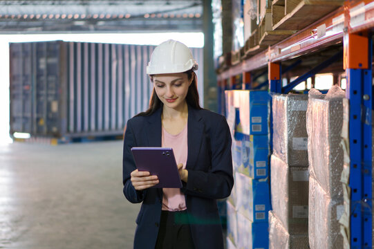Young business woman in suit and safety helmet review inventory record on tablet computer. Stand among stacked boxes in a busy storage area. Working in container stuffing and cargo securing service.