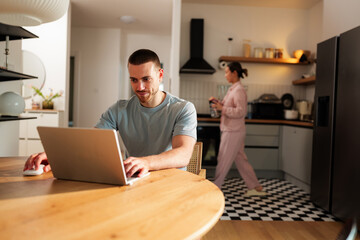 Young man concentrating on his laptop, working remotely from the dining table, while his wife prepares breakfast in the background in their modern kitchen