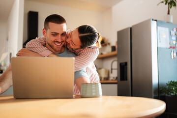 Young couple embracing while working on a laptop in a modern kitchen, enjoying each other's company during a cozy work from home day filled with love and connection