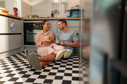 Young couple sitting on the kitchen floor, wearing pajamas and slippers, enjoying a cup of coffee together in the morning, chatting and bonding in a cozy domestic setting