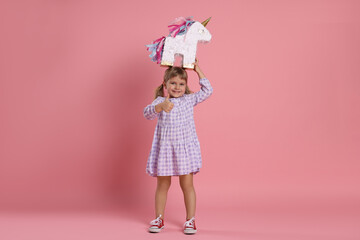 Happy girl with unicorn shaped pinata showing thumbs up on pink background