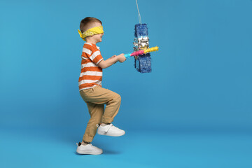 Happy boy with tied eyes breaking rocket shaped pinata on light blue background © New Africa