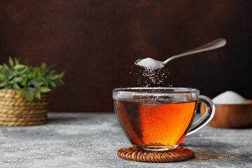 White sugar falling from teaspoon into glass cup of hot tea placed on coaster against dark brown background