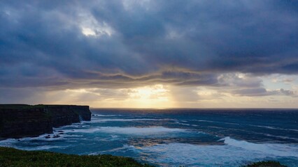 Serene view of sun setting over ocean, with sky full of clouds and cliff's edge in background.