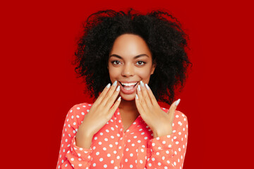 Surprised African American woman with natural curly hair, wearing a coral pink polka dot shirt, showing off her manicured nails against a bold red background..