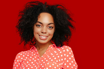 Smiling African American woman with natural curly hair, wearing a coral pink polka dot shirt, posing against a bold red background.
